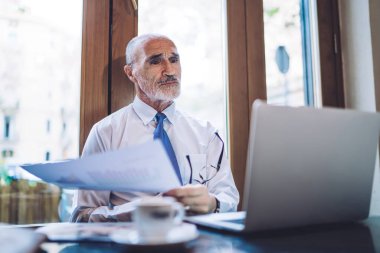 Gözlüklü yaşlı CEO, pencerenin yanındaki kahve fincanıyla masada otururken dizüstü bilgisayara bakıyor ve kağıt çarşaf tutuyor.