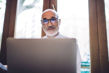 Beyaz gömlekli, mavi kravatlı yaşlı, kel, sakallı yönetici. Pencerenin yanındaki bulanık arka planda bilgisayar kullanıyor.