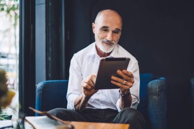 Gülümseyen, kel, resmi kıyafetler giyen, modern tableti zevkle karıştıran, Cafe 'deki koltukta oturan yaşlı adama sevindim.