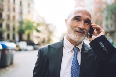 Gülümseyen, takım elbiseli, beyaz sakallı, sokakta yürürken telefonla konuşan, bulanık binalara bakan yaşlı, yakışıklı bir erkek. 