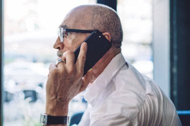 Beyaz gömlekli, gözlüklü, kol saati takmış akıllı telefondan konuşan ve pencereden arka plandaki güneş ışığına bakan sakallı yaşlı adamın yan görüntüsü.