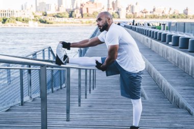 Kendine güvenen Afrikalı Amerikalı sporcunun sabah saatlerinde demir çitlerin üzerinde bacaklarını esneterek arka plandaki deniz manzarasına bakması.