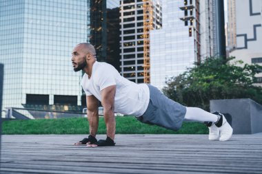 Spor giyimli, kaslı, kel, sakallı siyahi bir adamın modern şehir merkezinin caddesinde antrenman yaparken arka planda gökdelenlerle birlikte şınav çekerken yan görüntüsü.