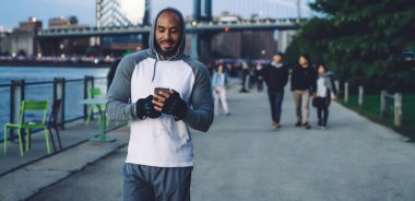 Gülümseyen atletik Afro-Amerikalı adam, rahat spor kıyafetleri içinde. Akşamları Megapolis 'teki rıhtımda dinlenirken akıllı telefondan mesaj atıyor.