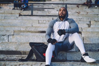 Genç Afrikalı Amerikalı atletik erkek taş merdivenlerde oturuyor ve kulaklıkla müzik dinliyor.