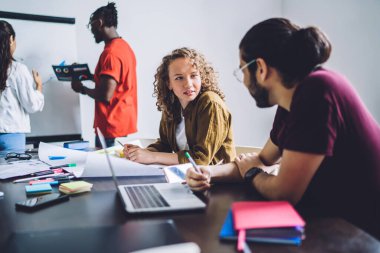 Kıvırcık kadın ve adamın masada dizüstü bilgisayarı ve beyaz tahta yakınında çeşitli grup arkadaşlarıyla sohbet edişinin yan görüntüsü.
