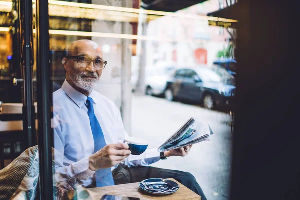 穿着短发的白衬衫 咖啡杯和报纸的老年男子坐在窗前 望着路边的街道 — 图库照片