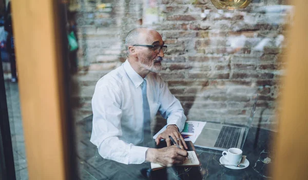 坐在咖啡店边浏览笔记本电脑时 透过玻璃窗观看戴眼镜 身穿正式衣服 在笔记本上记笔记的高年级秃头男性 — 图库照片