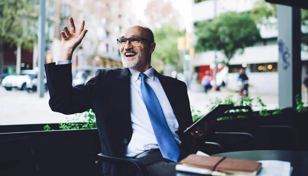 Delighted bald gray bearded senior man with eyeglasses in suit and blue tie sitting outside cafe and waving hand while using tablet