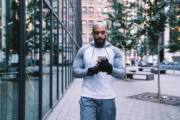 严重秃头的非裔美国男性 穿着运动服 站在现代建筑附近的街上 一边用智能手机一边听音乐 — 图库照片