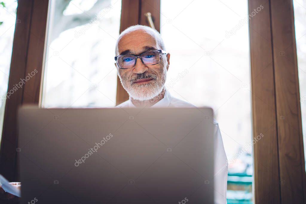 Senior bald gray bearded executive in white shirt and blue tie with glasses using pc near window on blurred background