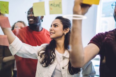 Yeni proje fikri üzerinde çalışırken yapışkan notlar düzenleyen modern kadın ve erkeklerin sırıttığı cam duvarın ötesine bak 