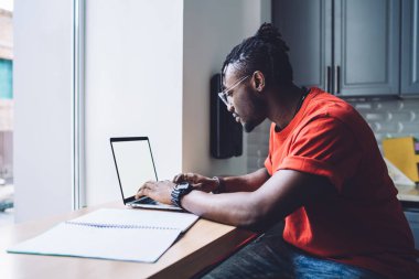 Modern Afro-Amerikan gözlüklü adamın yan görüntüsü. Tezgahta boş ekranlı dizüstü bilgisayarda oturuyor.