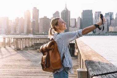 Amerikan kaçamağının tadını çıkaran turistik sırt çantalı neşeli kadın gezginin yandan görünüşü geçmişe dönük fotoğraf makinesi, mutluluk konsepti ve ABD tatilleri ile özçekim fotoğrafı çekmek için poz veriyor.