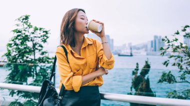 Sarı gömlekli ve sırt çantalı hoş bir Asyalı kadın Hong Kong rıhtımında durmuş kahve içiyor.