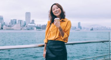 Yarım boy, modaya uygun giyinmiş, Hong Kong manzarasında kahve eşliğinde poz veren neşeli kadın portresi, Çin 'de tatil tatili yapmaktan mutlu komik gülümseyen kadın turist.