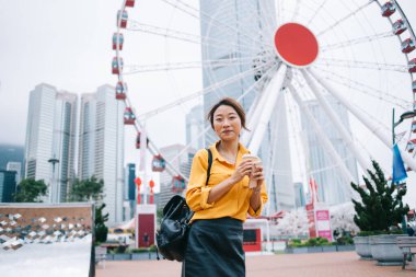 Hong Kong caddesinde dönme dolaba gülümserken, elinde kahve fincanı tutan sıradan giyinmiş, mutlu, genç bir kadın. 