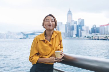 Şık giyinmiş, elinde kahve fincanı ve cep telefonuyla Hong Kong rıhtımına yaslanırken kameraya gülümseyen neşeli genç Asyalı kadın. 