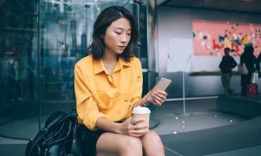 Modern elbiseli düşünceli bir Asyalı kadın sırt çantasıyla oturuyor ve Hong Kong 'da cep telefonuyla sörf yaparken kahve içiyor.