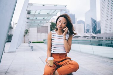 Plastik bardak ve bacak bacak üstüne atmış Asyalı güzel bir genç kadın telefonda konuşurken bankta oturuyor ve Hong Kong 'un arka planındaki kameraya bakıyordu.