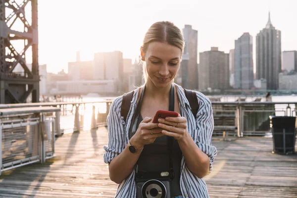 Happy Caucasian amateur photographer with instant camera on neck typing text article about American holidays in New York for sharing to social network and attracting new followers to own web page