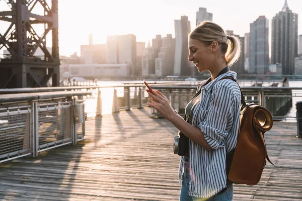 携帯電話を介してフォロワーとのネットワーキングやメッセージングながら 幸せな女性旅行ブロガーのサイドビューソーシャルパブリケーションのためのテキストを入力し 米国のウェブサイトを閲覧するために4Gの無線を使用して陽気な女性 — ストック写真