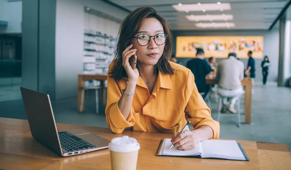 Привлекательная Азиатская Женщина Очках Делающая Заметки Разговаривающая Телефону Перед Ноутбуком — стоковое фото