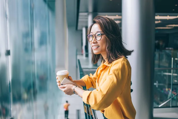 明るい肯定的なアジアの女性の側面図ガラスとホットドリンクと携帯電話で黄色のシャツ手すりにもたれて離れて見て 香港で屋内で考える — ストック写真