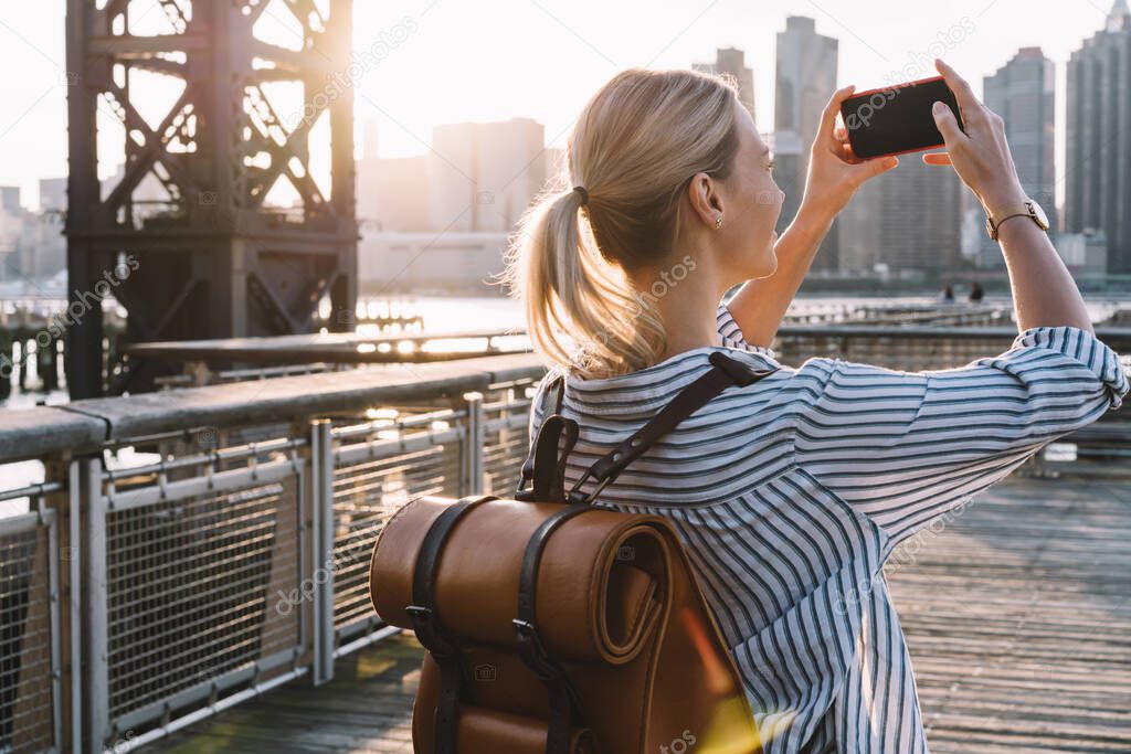 Back view of millennial woman in casual wear shooting video of New York landscape using smartphone main camera for clicking pictures, female tourist testing cellphone while taking photos of city