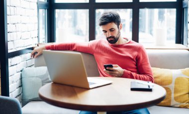 Milenyum erkek blogcu profili akıllı telefon ağlarında 4G internet bağlantısıyla modern dizüstü bilgisayarlı şık bir kafede dinleniyor cep telefonuyla sohbet eden adam