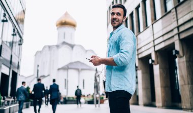 Havalı tişört giymiş, şık giyinmiş, gülümseyen ve akıllı telefoncunun sesli şarkısını dinleyen kulaklıklı mutlu erkek melodi sanatçısı boş zamanlarında müzik podcast 'i dinliyor.