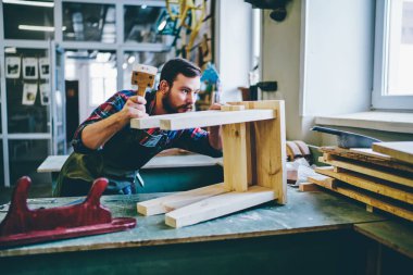 Ahşap teçhizatla mühendislik yapan serbest çalışan restorasyon imalat sahibi, önlüklü beyaz adam, çalışma sırasında dekoratif el yapımı sandalye geliştirmeye odaklanmış.