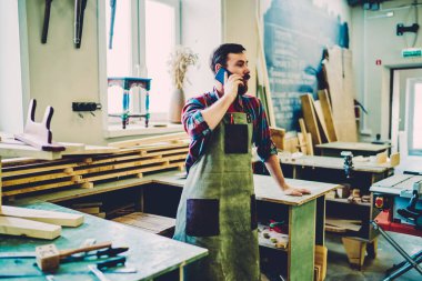 Beyaz önlüklü bir zanaatkar, ahşap imalat fabrikasının teslimat zamanını tartışmak için önemli bir cep telefonu konuşması yapıyor. Çevrimiçi iletişim konsepti.