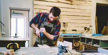 Kendi atölyesinde mobilya için ahşap kalasla çalışırken aprondaki usta zanaatkar, sakallı adam marangoz tamir ve onarım için modern elektrikli teçhizat kullanıyor.