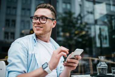 Millennial yakışıklı adam mavi gömlekli ve gözlüklü cep telefonu kullanıyor ve sokak kafesi terasında otururken gülümsüyor.