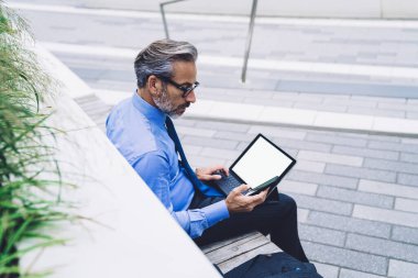 İş yerinde sakallı, olgun bir erkeğin oturduğu ve elinde boş dizli tabletle New York caddesinde telefonu kullandığının yan görüntüsü.