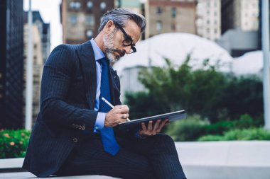 New York sokaklarında bankta otururken Stylus 'la tablet üzerinde çalışırken bacak bacak bacak üstüne atmış orta yaşlı işadamının yan görüntüsü.