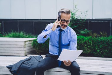 New York şehir merkezindeki bankta gözlüklü orta yaşlı bir işadamı iş belgelerini tutuyor ve telefonla konuşuyor.