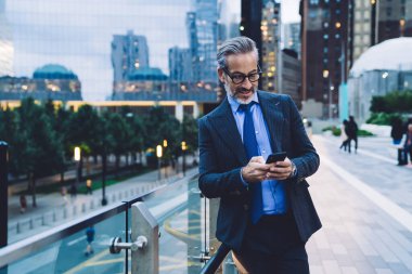 Yakışıklı, gri saçlı, modaya uygun saç kesimli, siyah takım elbiseli, mavi kravatlı, akıllı telefondan mesaj atan, cam çitlere yaslanan, New York caddesinde bulanık arka planda oturan yakışıklı bir iş adamı.