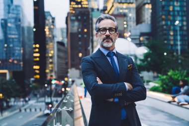 Yakışıklı, orta yaşlı, gözlüklü, takım elbiseli, kolları çaprazlanmış, cam çitin yanında duran New York caddesine karşı.