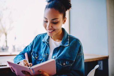 Kot ceketli mutlu bayan öğrenci üniversite dersleri için fikirler yazıyor eğitim kitaplarında yazıyor, başarılı Afrikalı kadın gazeteci toplantılar ve gelecekteki röportajlar hakkında bilgi alıyor