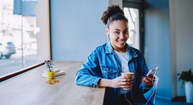 Elektronik kulaklıklı mutlu koyu tenli hippi kız portresi yüksek sesli müzik dinleyen, müzik listesinden hoşlanan ve kahve molası sırasında kameraya bakan genç ve neşeli kadın.