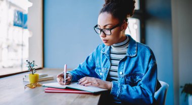 Ciddi koyu tenli kadın blog yazarı kafeterya masasında oturuyor ve sosyal ağda bir sonraki yayın için fikirler yazıyor, zeki öğrenci sınav notlarına ders kitabında hazırlanıyor