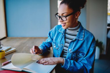 Kafeteryada öğrenim gören mutlu kız öğrenci eğitim kitabında proje için bilgi arıyor. Gözlük takan neşeli Afrikalı hippi kız not defterine bakıyor.