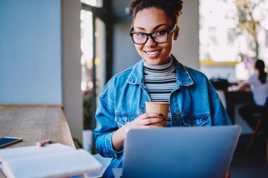 Kampüste öğrenimden ve kafeinden keyif alan yetenekli kız öğrencinin portresi, bilgisayarda çalışmak için başvuru kurulumunu beklerken kameraya bakan gülümseyen kadın.