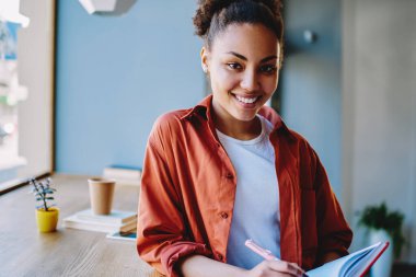 Zengin Afrikalı kadın yazarın yarım boy portresi. Elinde ders kitabı, boş zamanlarında kameraya bakıyor. Kahve dükkanında ders çalışmaktan zevk alan esmer tenli bir kadın.