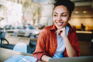 Gülümseyen kız öğrenci, ara verdiğimizde laptoptan öğrenmeyi düşünüyor. Pozitif esmer tenli kadın üniversite kampüsünde günübirlik giyinmiş.