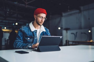 Genç stil odaklı sakallı erkek. Sıcak kıyafetler ve gözlüklü. Ofiste tabletle beyaz masada oturuyor.