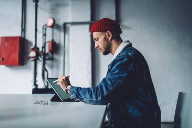 Tarz sahibi, sakallı, serbest çalışan erkek tasarımcı sıcak mavi kot ceket giyiyor ve ofisteki tablette şapka testi uygulaması çiziyor.