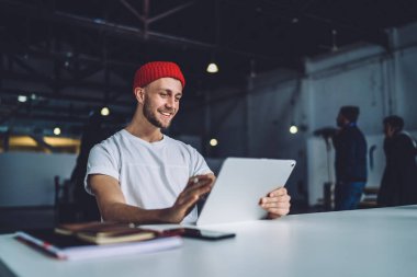 Beyaz tişörtlü neşeli, şık hippi adam modern garajda aynı masada oturuyor ve modern tablet aletlerini karıştırıyor.